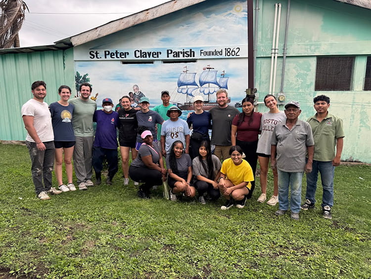 Fairfield students during their immersion trip to Belize, standing in a group in front of a 'St. Claver Parish' mural.