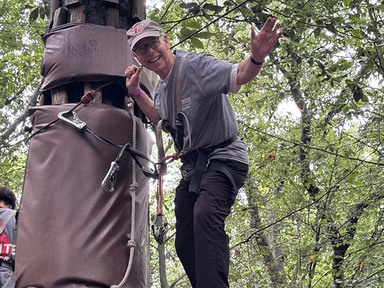 A smiling Father Savard in a gray shirt and safety harness waves while climbing beside a large tree trunk, with ropes and leafy branches around him.