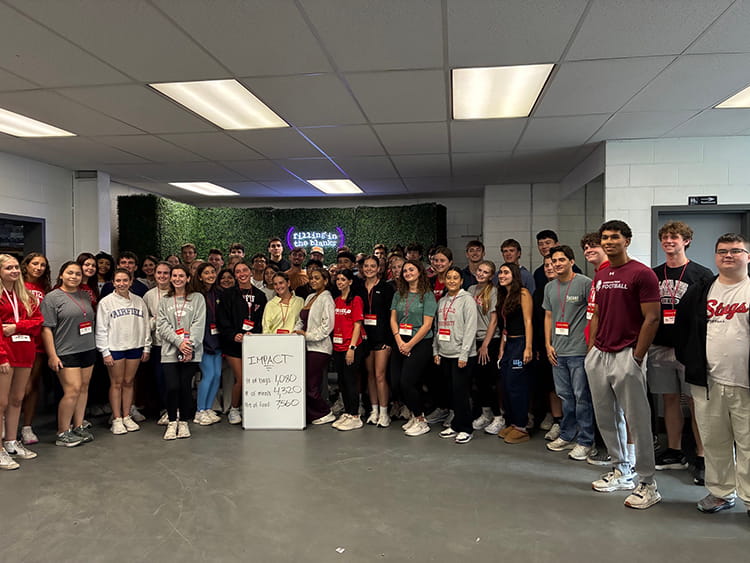 A large group of students and staff stand in rows in a classroom or conference room, smiling toward the camera. A presentation screen and whiteboard are behind them, creating a friendly, organized group-photo atmosphere.