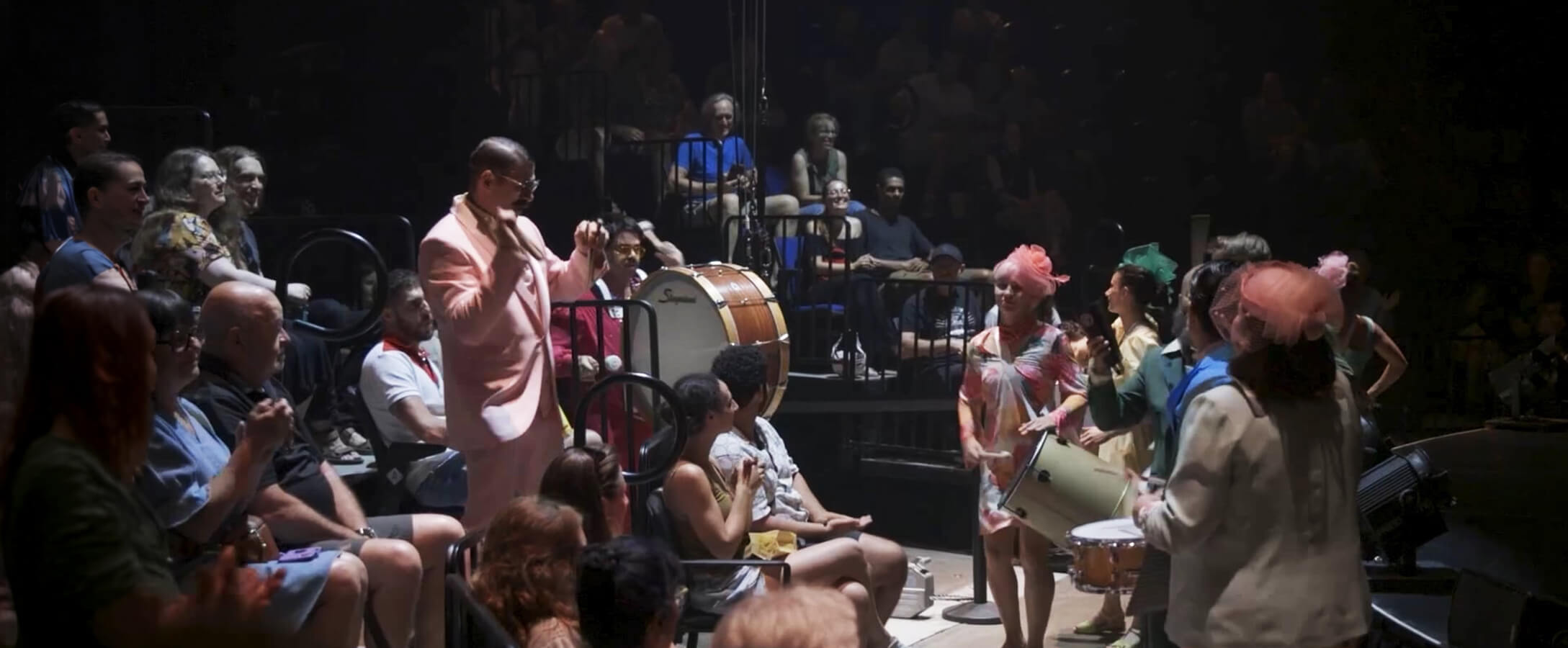 A vibrant performance scene features a band with colorful outfits and instruments playing among seated, applauding audience members, creating a lively atmosphere.