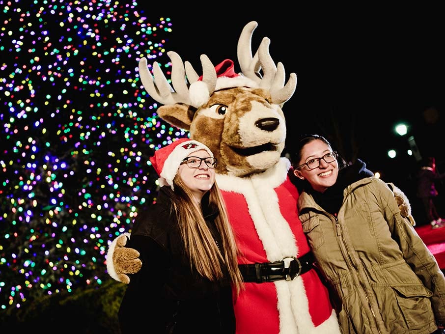 Students smile and pose with Fairfield's Lucas the Stag mascot in front of Fairfield's annually-decorated Christmas tree.
