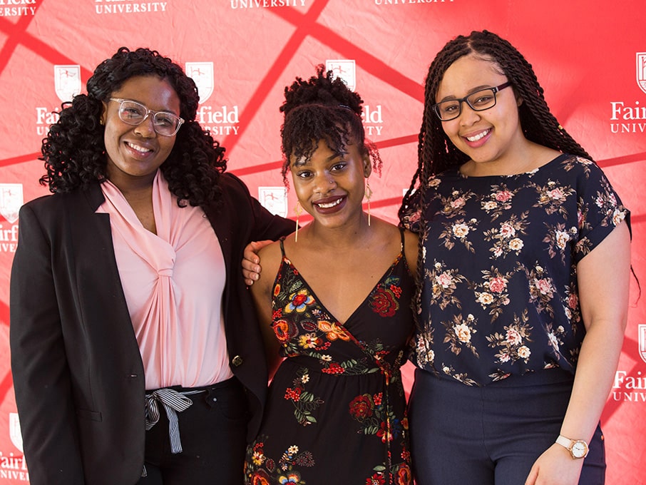 Fairfield students posing as a group of three against a Fairfield-branded backdrop at a campus event.