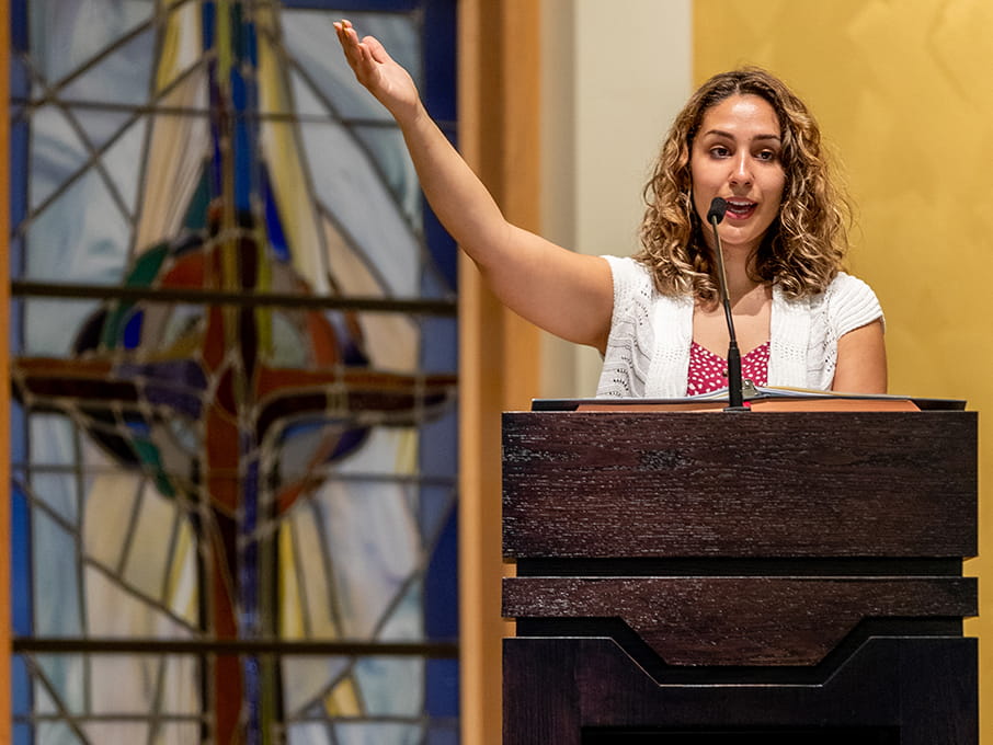 Curly-haired woman speaks into a microphone at a wooden podium, raising one arm energetically before a colorful stained-glass window.