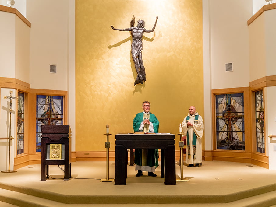 Catholic church altar with two priests in green and white vestments beneath a large crucifix, glowing gold backdrop, and stained-glass windows.