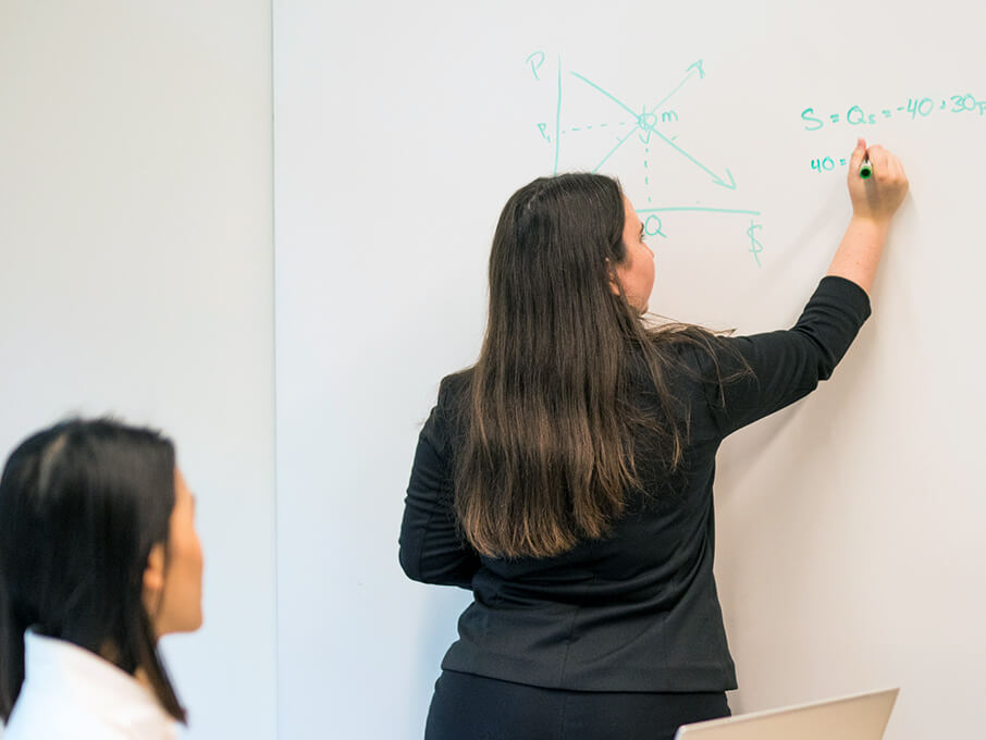 A woman writes geometric equations on a whiteboard while a colleague watches, creating an engaged, collaborative classroom or meeting atmosphere.