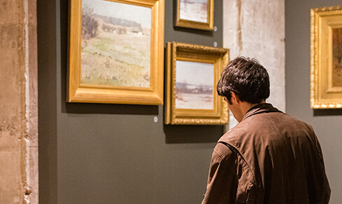 A museum visitor in a brown jacket studies framed landscape paintings on a dark gallery wall, creating a quiet, contemplative mood.