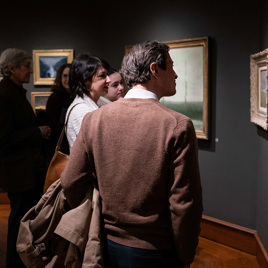 Museum-goers explore an art exhibit, intently looking at paintings and sharing thoughts on the artwork with one another.
