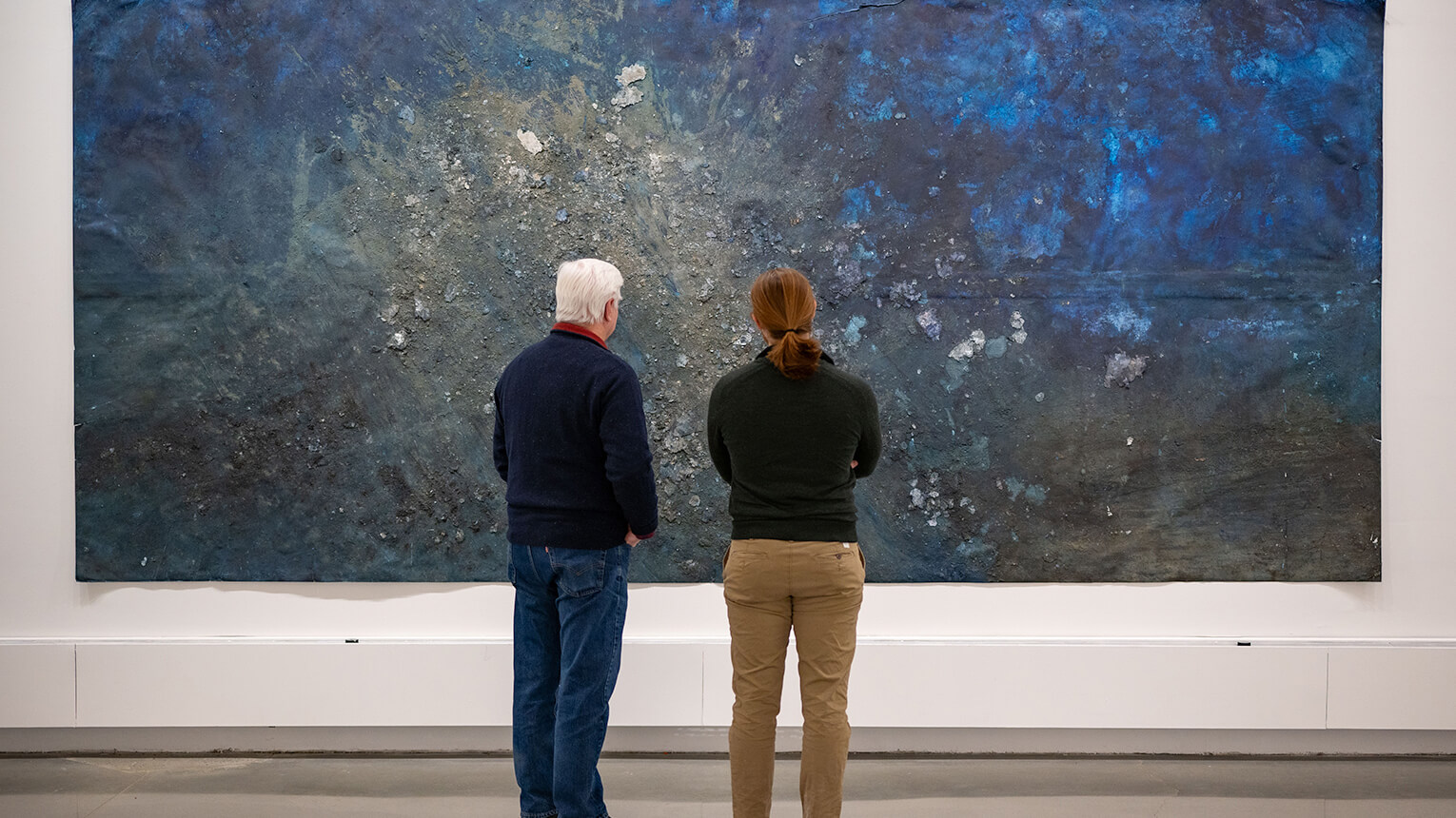 Two people stand in front of a large painting in an art museum, discussing its features and artistic style.  