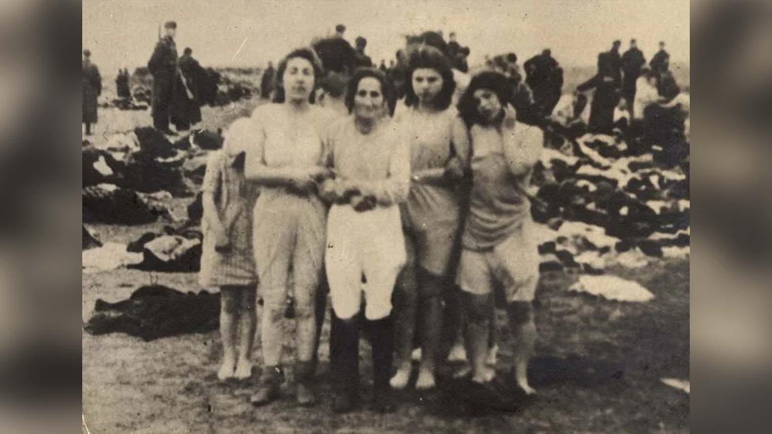 A group of women stand together outdoors, dressed in undergarments, with scattered clothing and people in the background. The image is in black and white.