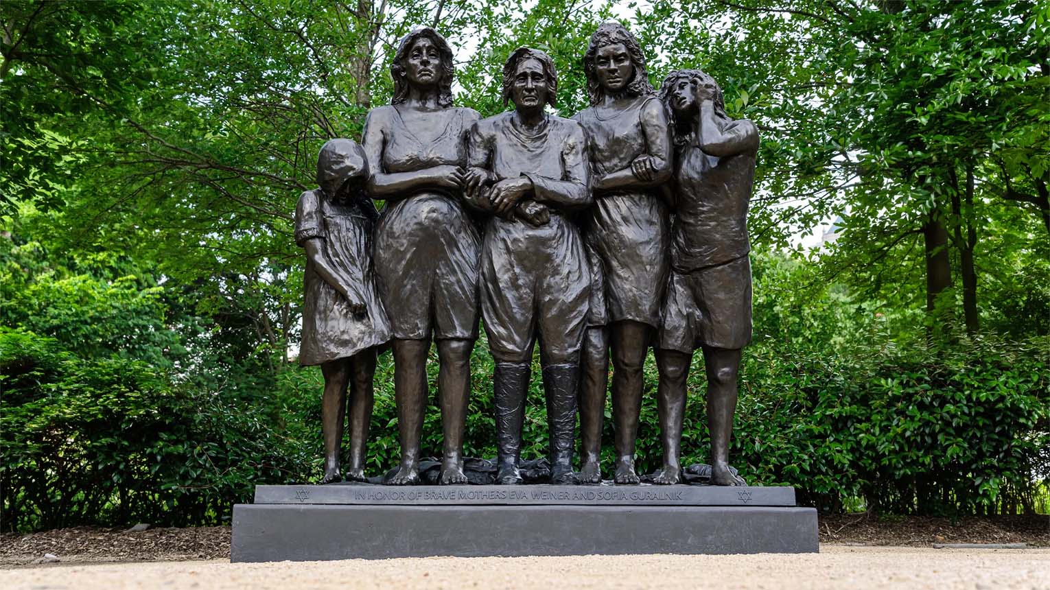 Bronze statue of five women and girls standing close together on a pedestal, surrounded by greenery; plaque reads, "In honor of brave mothers Eva, Wiena, and Sofia Subaliuk.