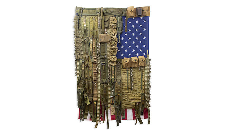 A U.S. flag covered with military gear, including belts and ammo pouches, hangs vertically. The arrangement evokes themes of war and patriotism.