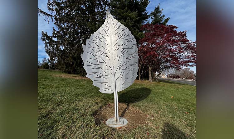 A large metal leaf sculpture stands prominently in a grassy area, showcasing its intricate design and shiny surface.