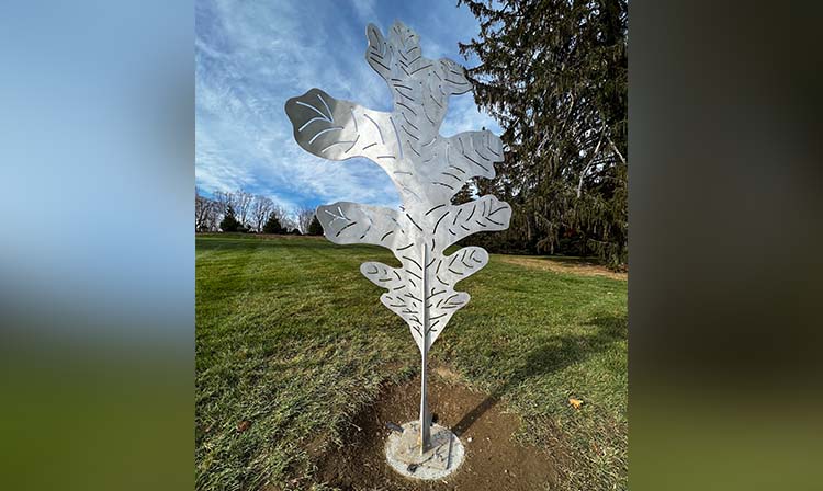 A metal sculpture of a tree stands tall in a grassy area, showcasing intricate branches and leaves.