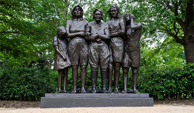 Bronze statue of five women and girls standing close together on a pedestal, surrounded by greenery; plaque reads, "In honor of brave mothers Eva, Wiena, and Sofia Subaliuk.