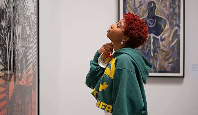 A woman stands in an art gallery, thoughtfully examining a piece of artwork displayed on the wall.