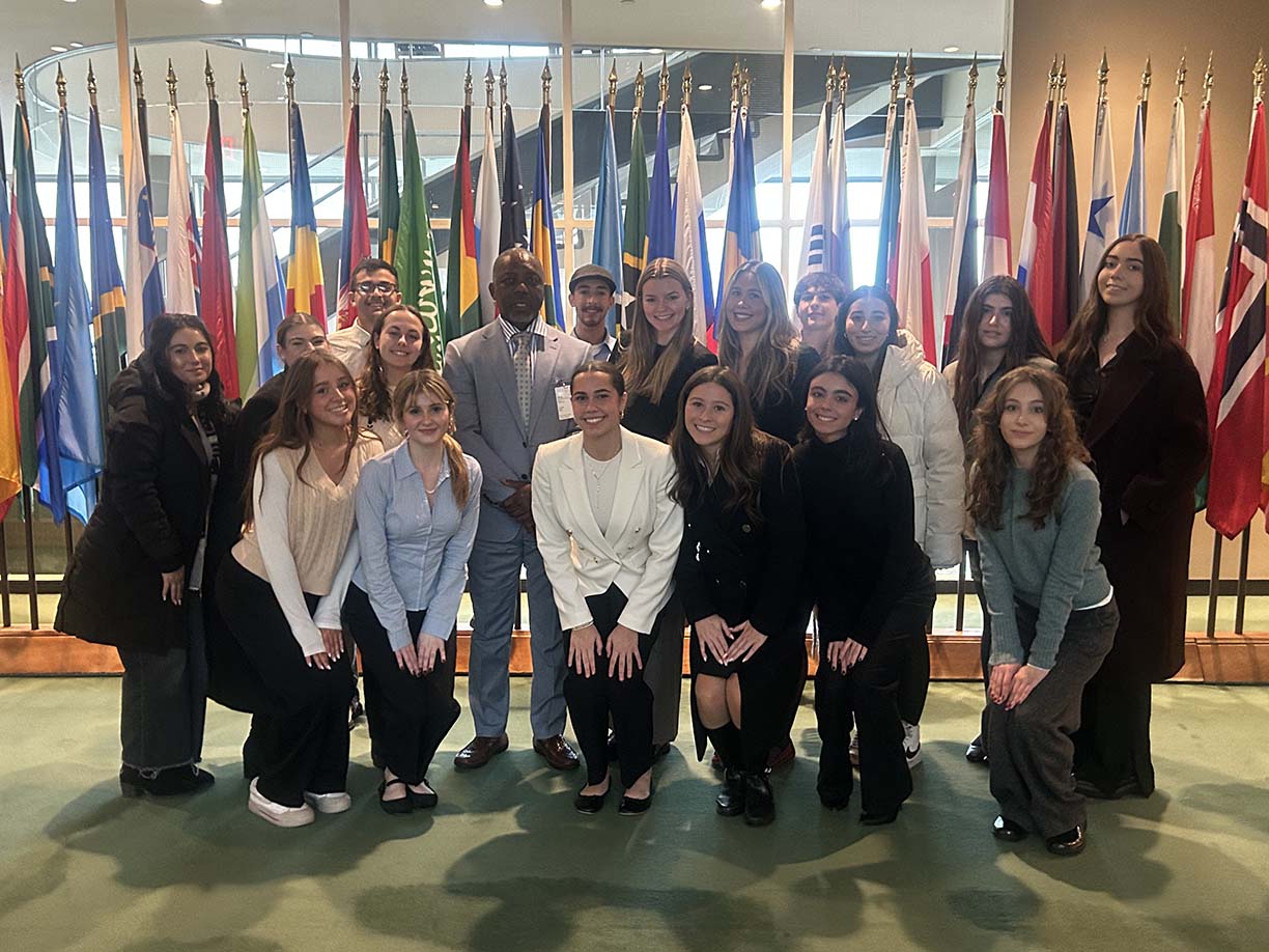 A diverse group of young adults and one man in a suit pose together in front of a display of various international flags inside a formal building.