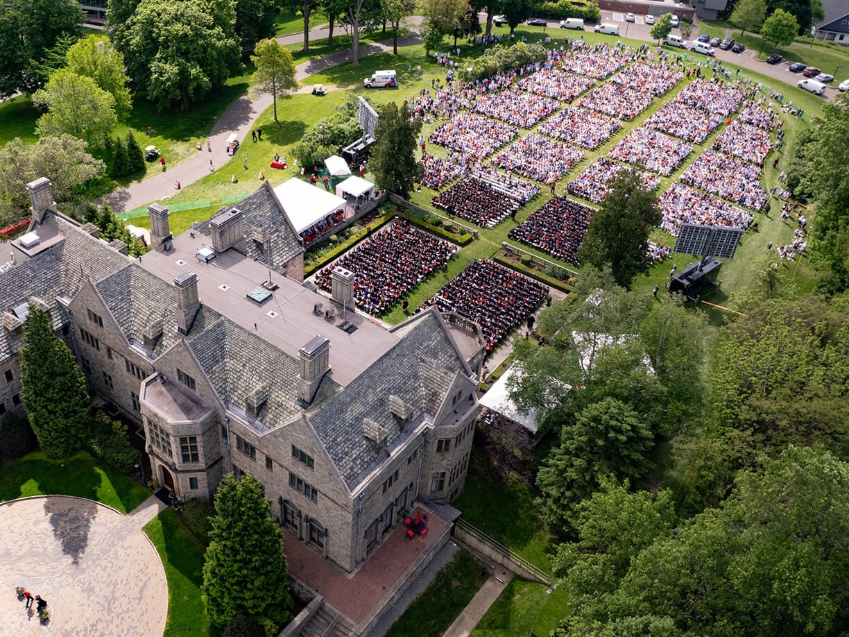 Aerial view of a Bellarmine Hall surrounded by lush trees. Nearby, a crowd gathers on a lawn for an outdoor event, suggesting a celebratory atmosphere.