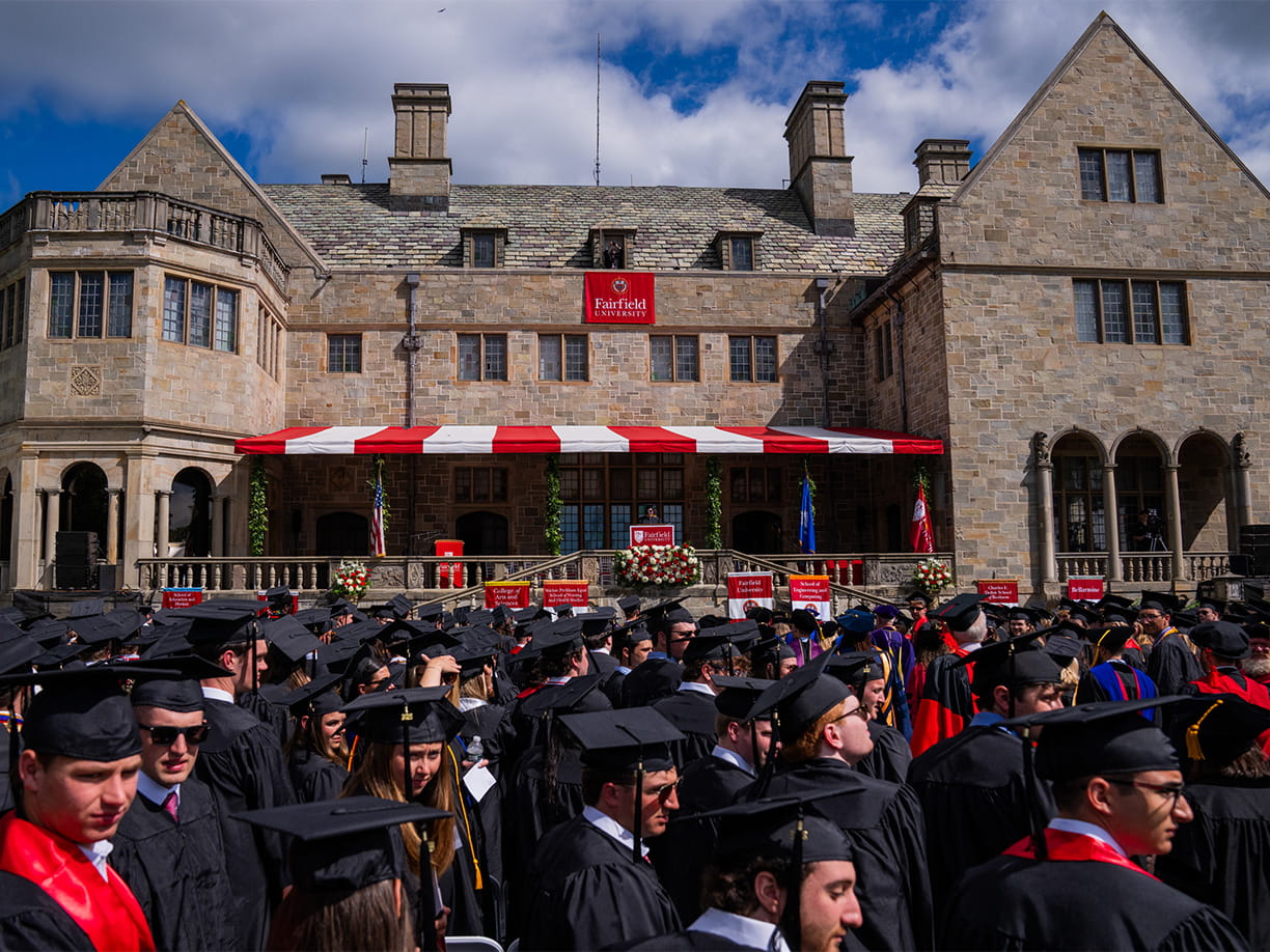 Students in caps and gowns lined up at their seats on a bright, sunny day as Commencement exercises begin.