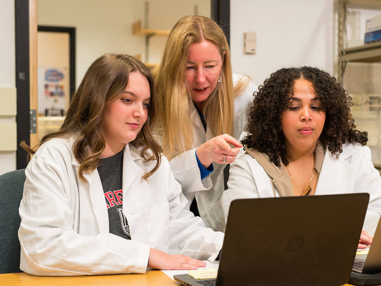 Shannon Harding, PhD and two other women in lab coats are engaged in a discussion around a laptop in a laboratory setting. One woman points at the screen, suggesting collaboration.