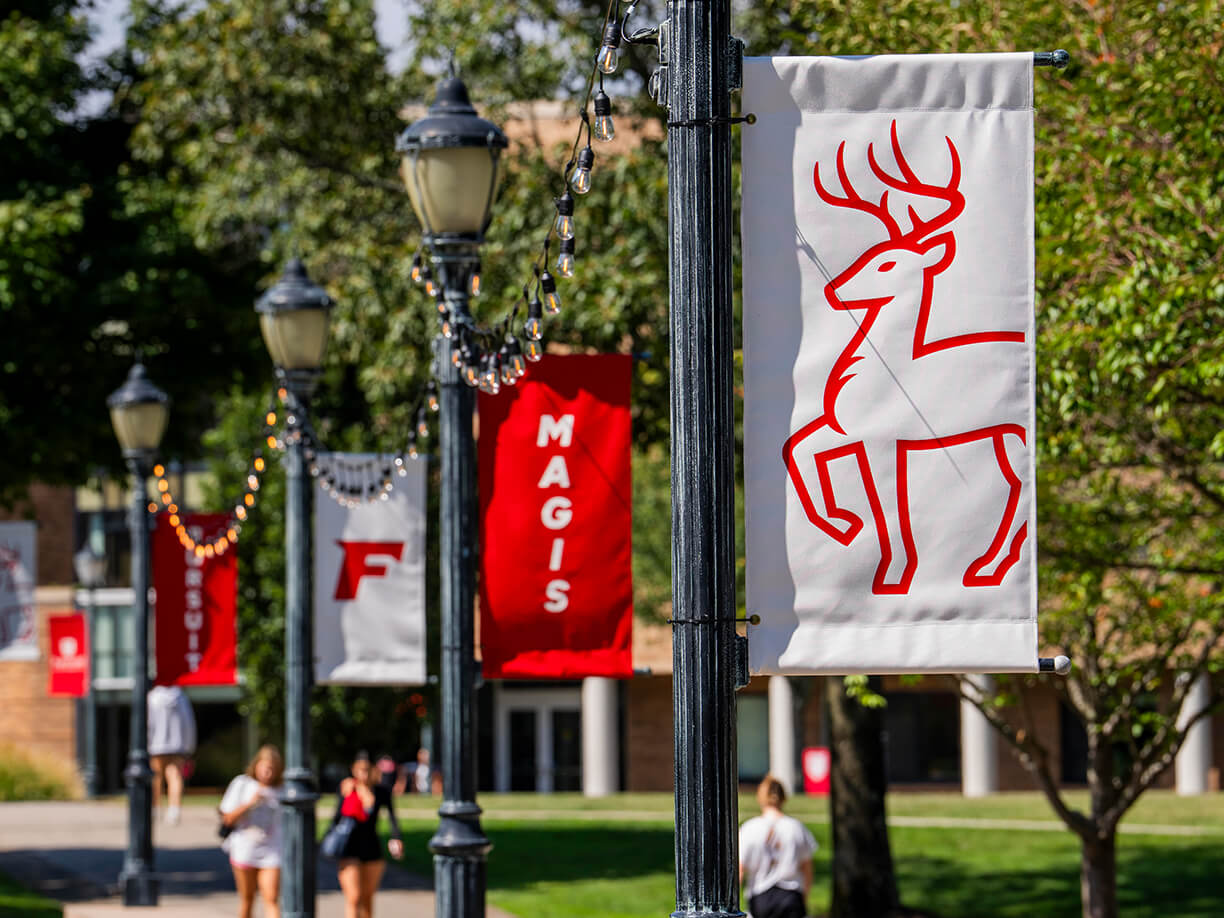 Fairfield University campus walkway with decorative light poles displaying red and white banners featuring the university stag logo and the word "Magis," surrounded by green trees on a sunny day.