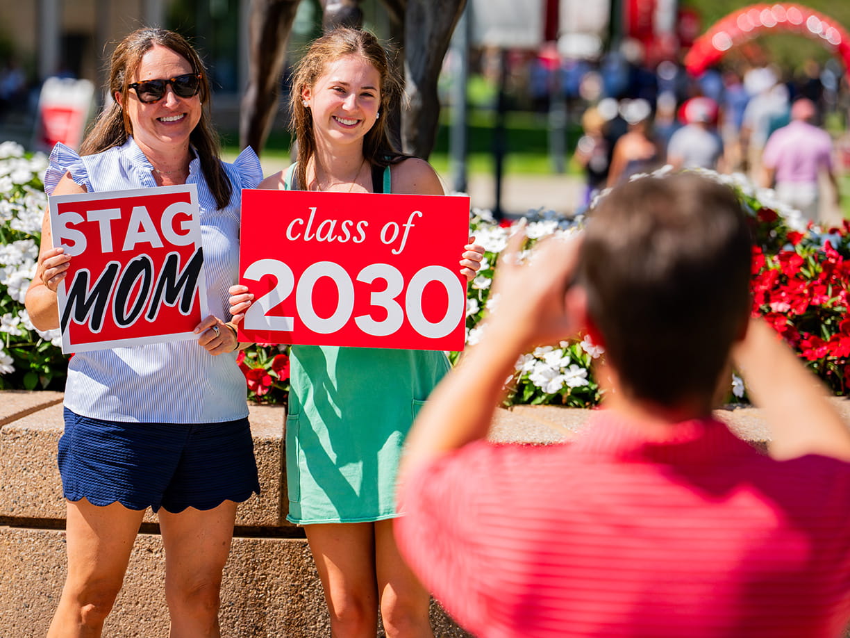 A first-year student holding up a written sign for 'Class of 2030' with her mom (with a written sign for 'Stag Mom') being photographed at move-in.
