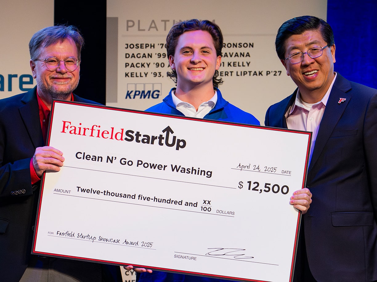 Male business sophomore with two faculty members smiling and holding a prop check for $12,500.