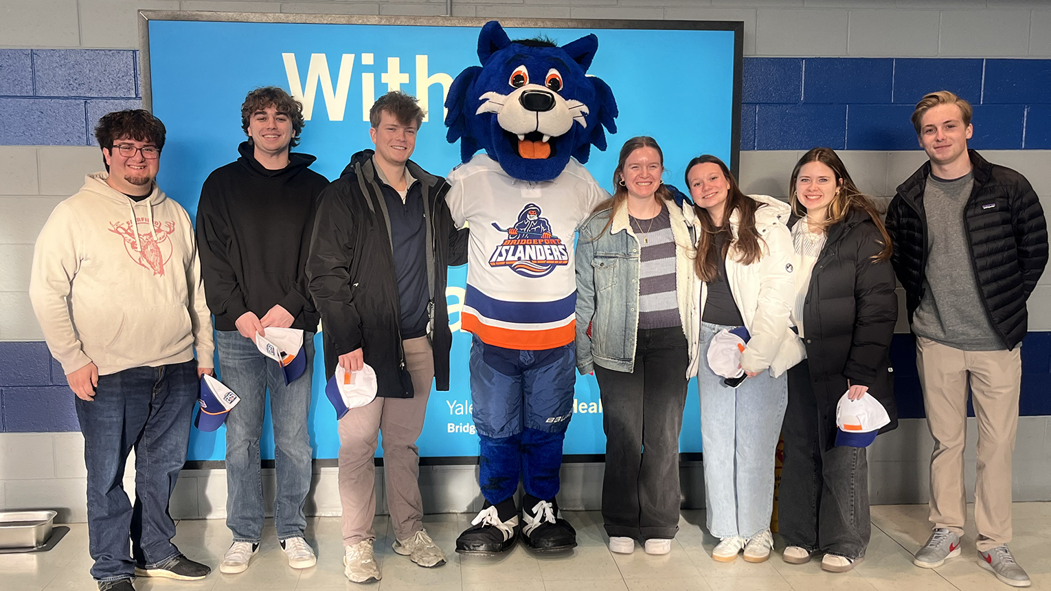 A group of smiling Fairfield students stands with a hockey team mascot in the center. The mascot wears a jersey, conveying a fun sports event mood.