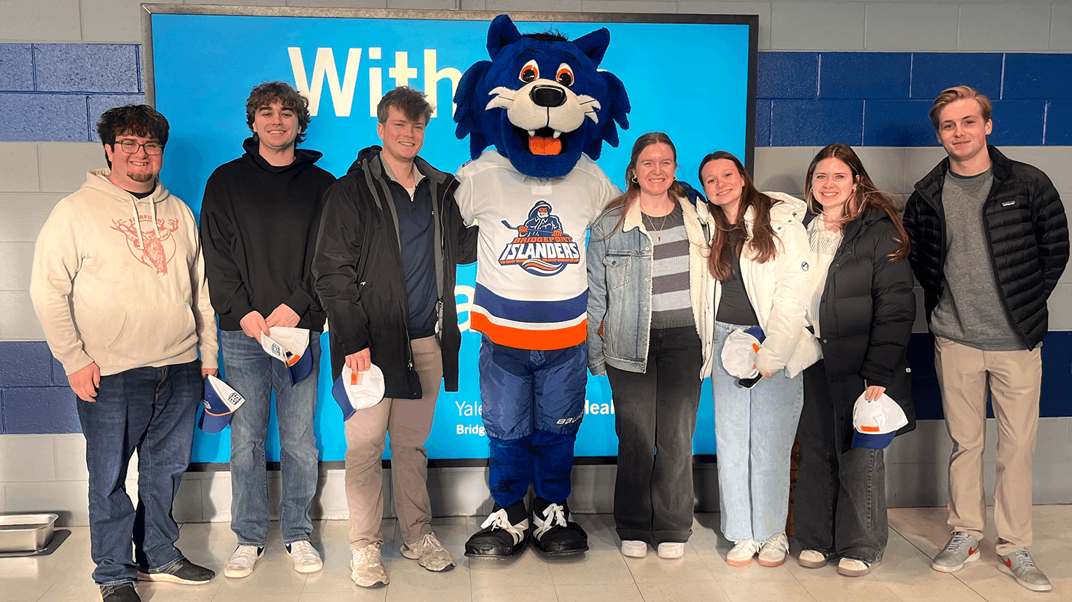 A group of smiling Fairfield students stands with a hockey team mascot in the center. The mascot wears a jersey, conveying a fun sports event mood.
