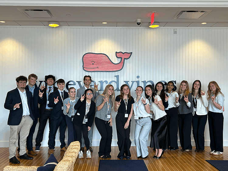 A group of Fairfield students pose in business attire, smiling and making "stags Up" hand signs in front of a wall with a pink whale logo and text that reads "Vineyard Vines."