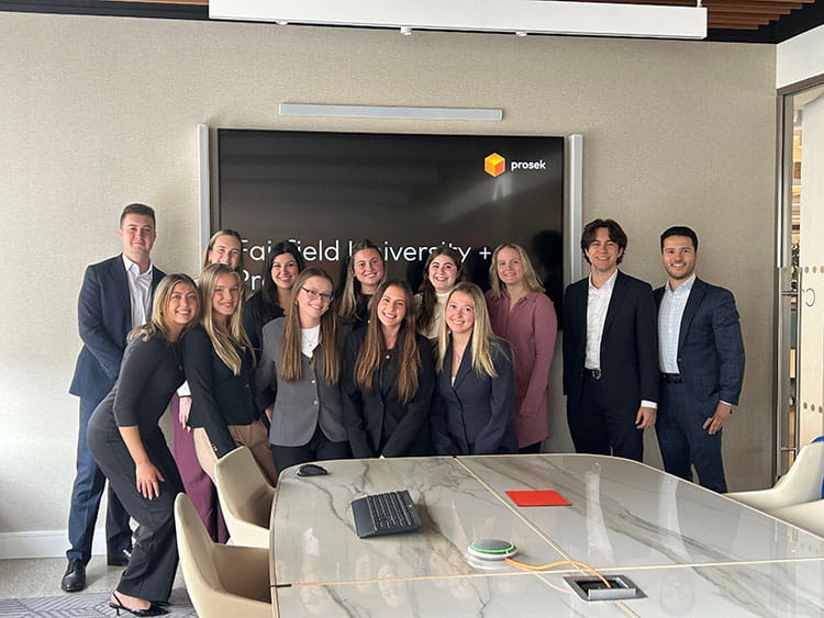 A group of Fairfield students dressed in professional attire smiles at the camera, standing in a conference room with a large screen displaying "Fairfield University + Prosek."