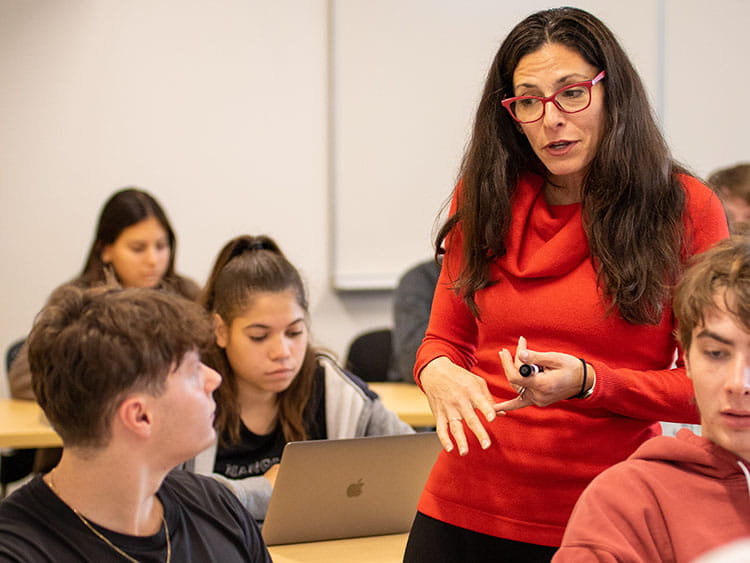 Professor Bloch engages with students in a classroom. Students, using laptops, appear attentive. The scene is focused and educational.