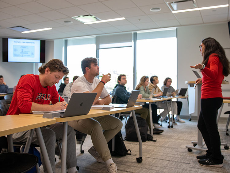 A classroom scene with students seated at desks with laptops, attentively listening to Professor Bloch in red who is speaking and gesturing at the front.