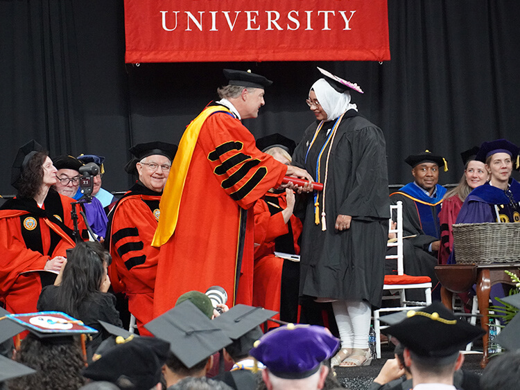 University graduation ceremony with a graduate in cap and gown receiving a diploma from a faculty member in academic regalia. Family and friends watch proudly.