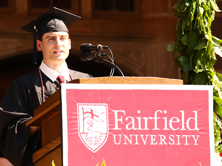 Tristan DeCrescenzo ’26 in cap and gown speaks at a podium adorned with a red Fairfield University banner. The atmosphere is formal and celebratory.