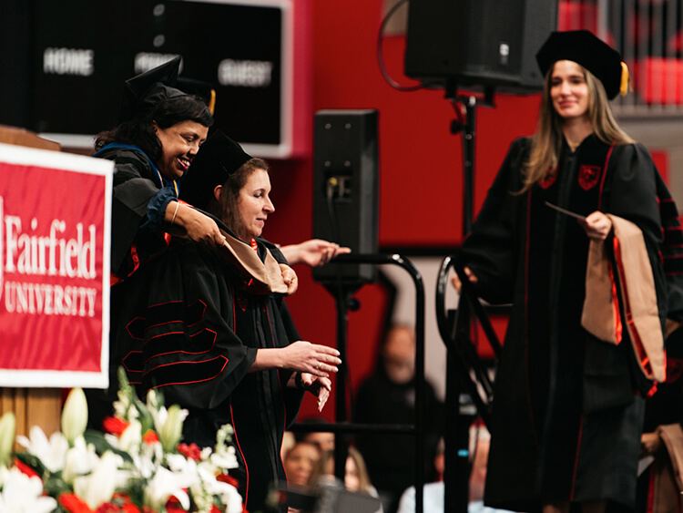 Two graduates in black robes being hooded by faculty members at a Fairfield University ceremony. The mood is celebratory, with flowers in the foreground.