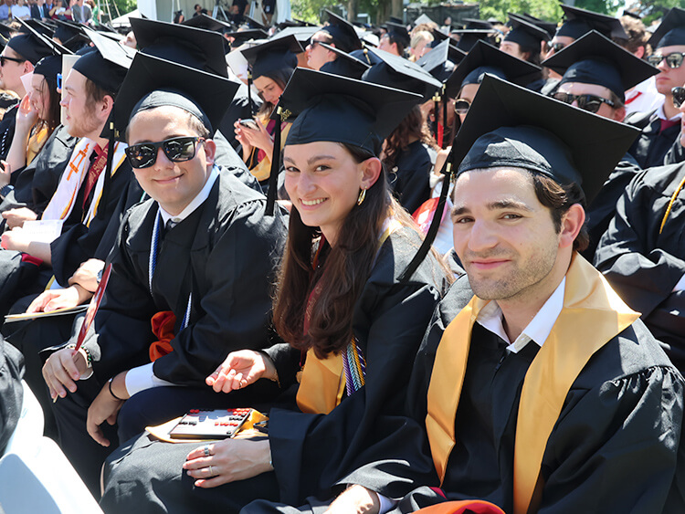 Graduating students in caps and gowns smile while seated outdoors. The sunny setting and their excitement convey a celebratory and joyful atmosphere.