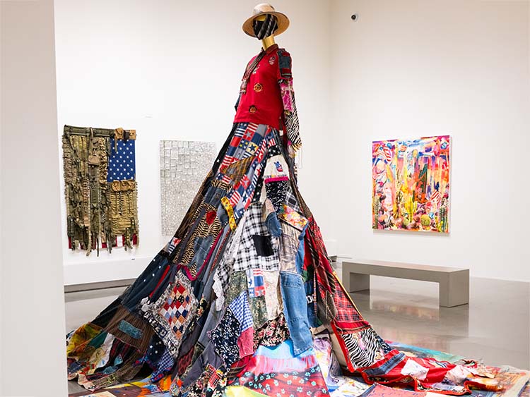 A tall mannequin in a patchwork dress dominates an art gallery. The colorful dress, titled “Freedom is Not Free?” and created by artist Maria de Los Angeles, flows outward, surrounded by abstract paintings on the walls, creating a vibrant, eclectic atmosphere.