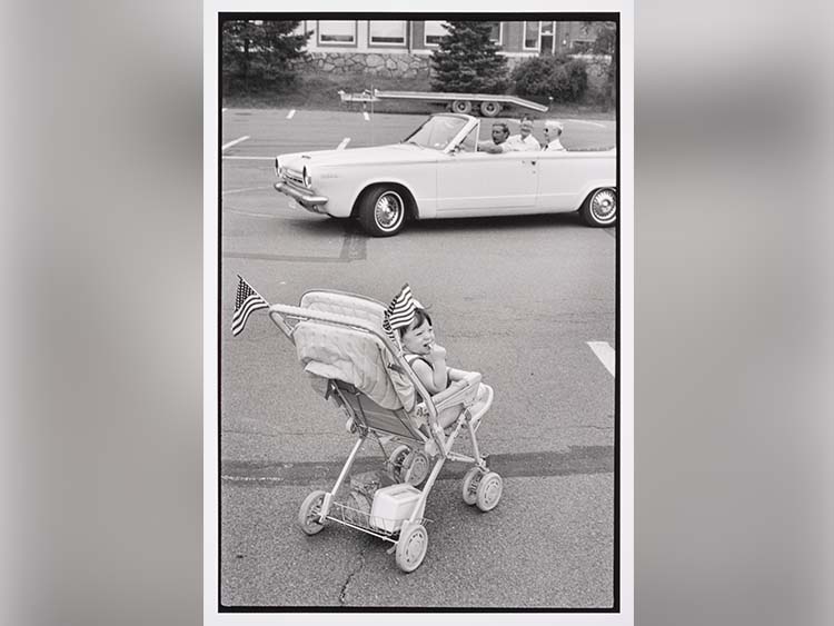 A black and white photograph by Leonard Freed titled “A baby sits in a stroller adorned with American flags” depicting a child in a stroller adorned with American flags watching a vintage convertible with three passengers drive by in a parking lot, evoking nostalgia.