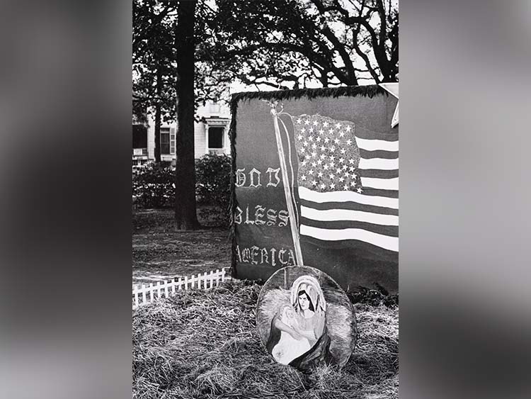 A black and white photograph by Leonard Freed titled “God Bless America—sign in private garden in South Carolina” depicting an American flag mural with "God Bless America" text, near a painting of a woman and child. A small white picket fence borders a straw-covered yard.