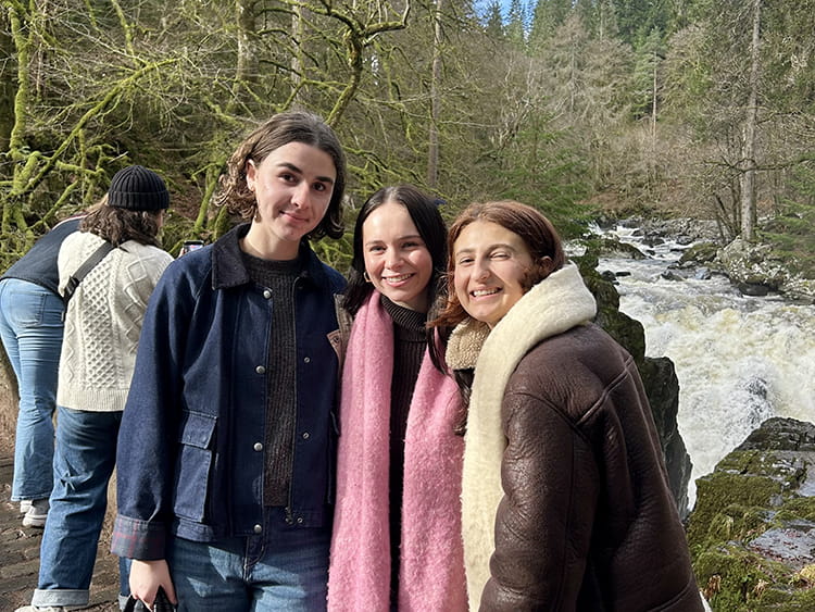 Three friends smile beside a rushing woodland waterfall: a long-haired person in a dark coat, a person in a pink scarf, and a person in a brown coat.