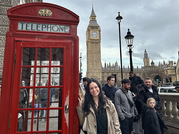 A smiling tourist stands beside a red British telephone booth as crowds cross Westminster Bridge, with Big Ben rising behind them under cloudy skies.