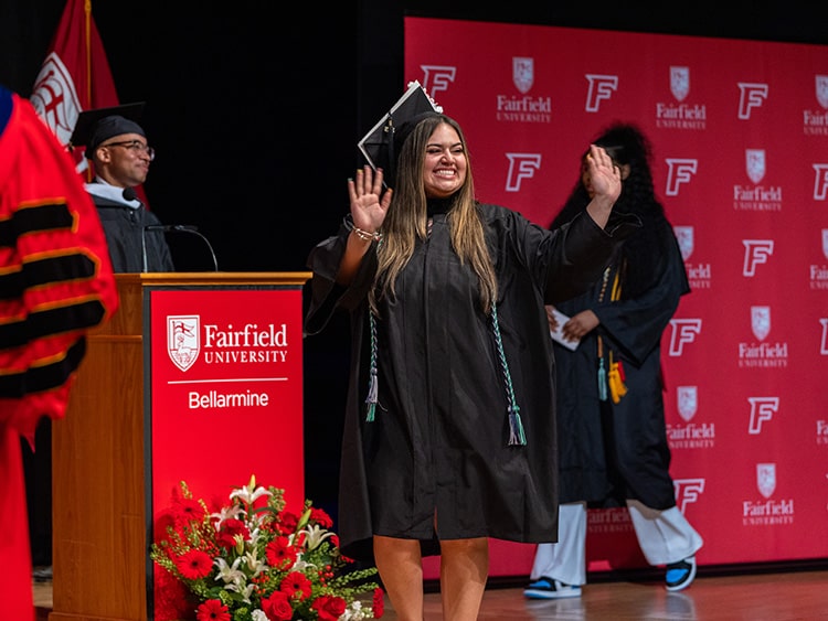 Female Fairfield Bellarmine student smiling and waving at the crowd as she exits the stage with her diploma in hand.