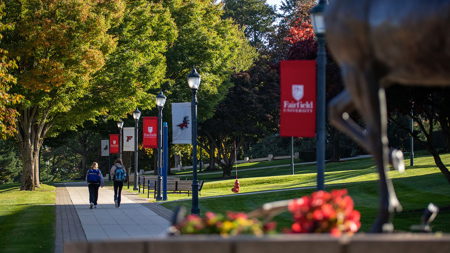 Two people walk on a path on campus next to a stag statue, with trees and grass framing the scene.