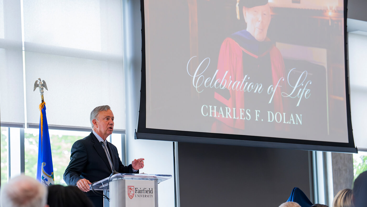A speaker stands at a podium, addressing an audience with a large screen presentation in the background.