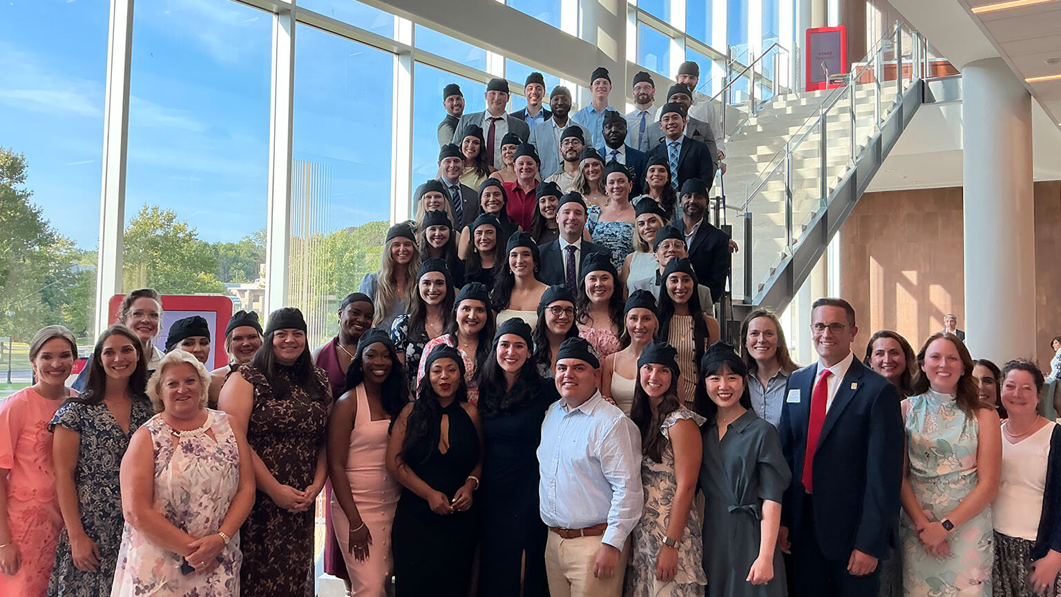 A cheerful group of students posing for a picture in front of a spacious window, allowing sunlight to illuminate the scene.