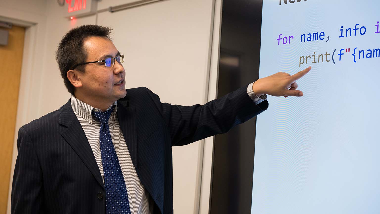 Image of a man in a suit and tie pointing at a screen, likely presenting information to an audience.