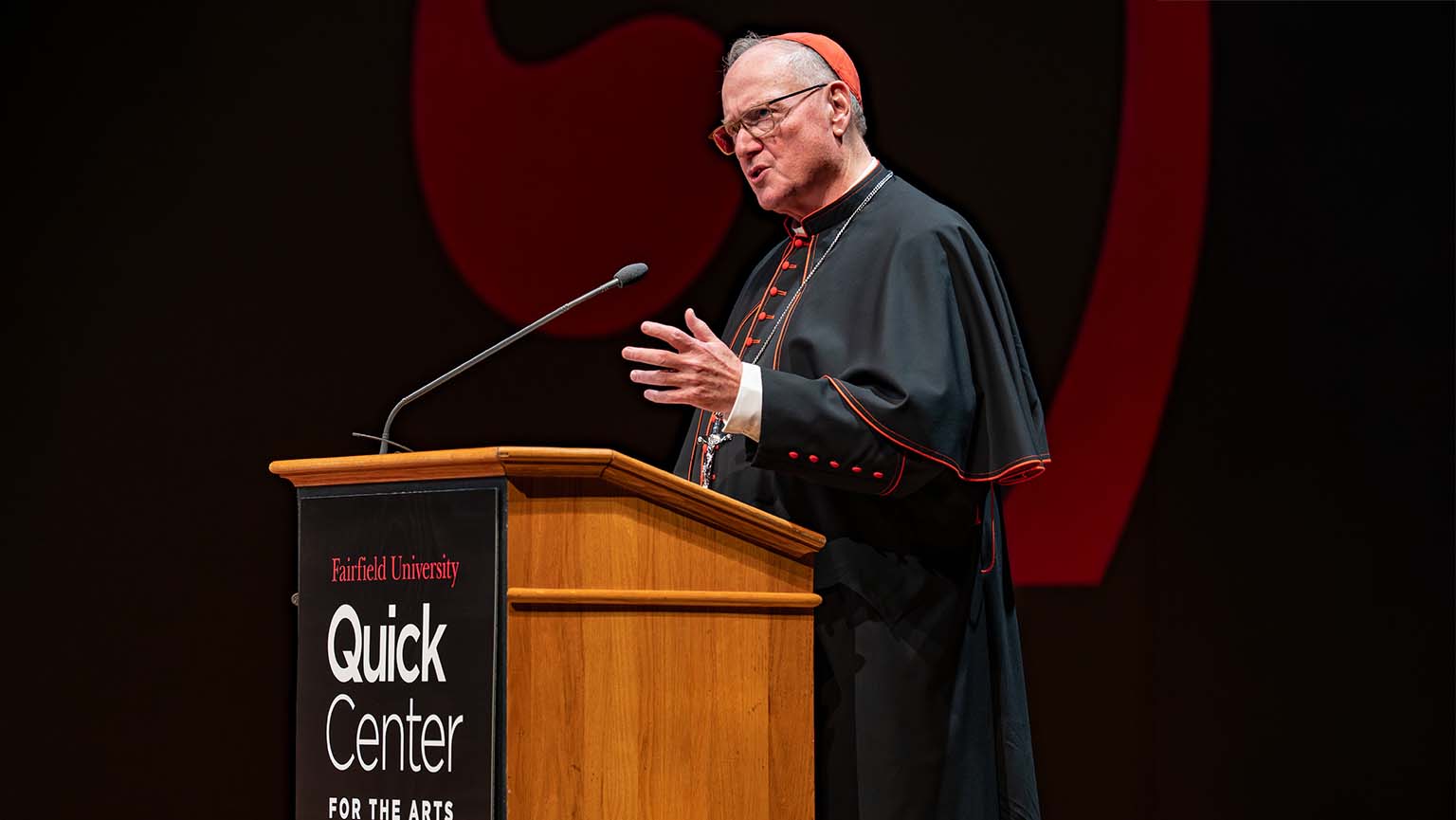 Image of Cardinal Dolan speaking at a podium on stage, engaging the audience with his message.