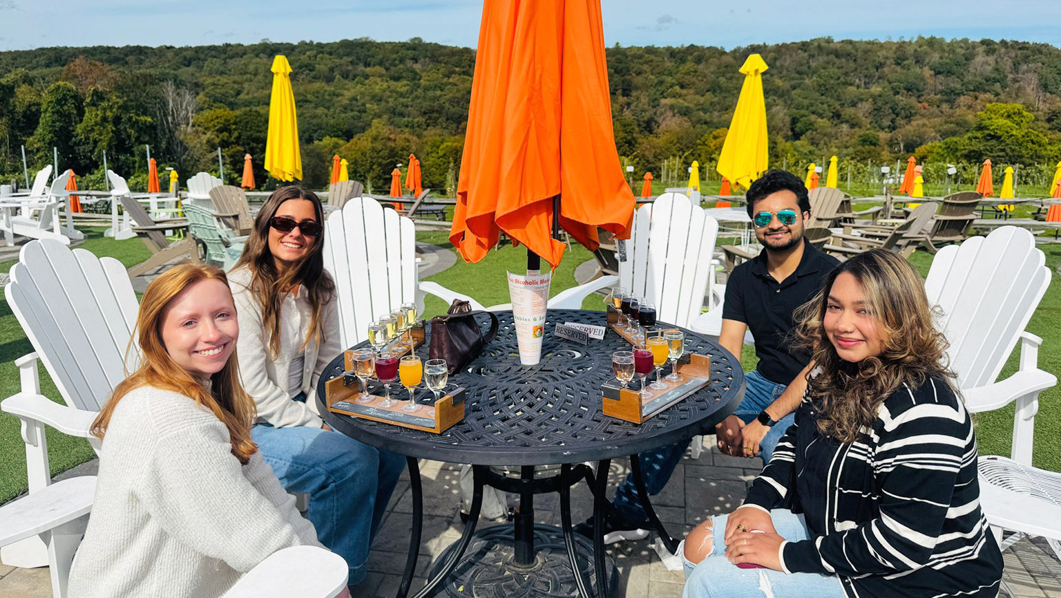 Four people at a table with an umbrella, engaged in conversation in a pleasant outdoor environment.