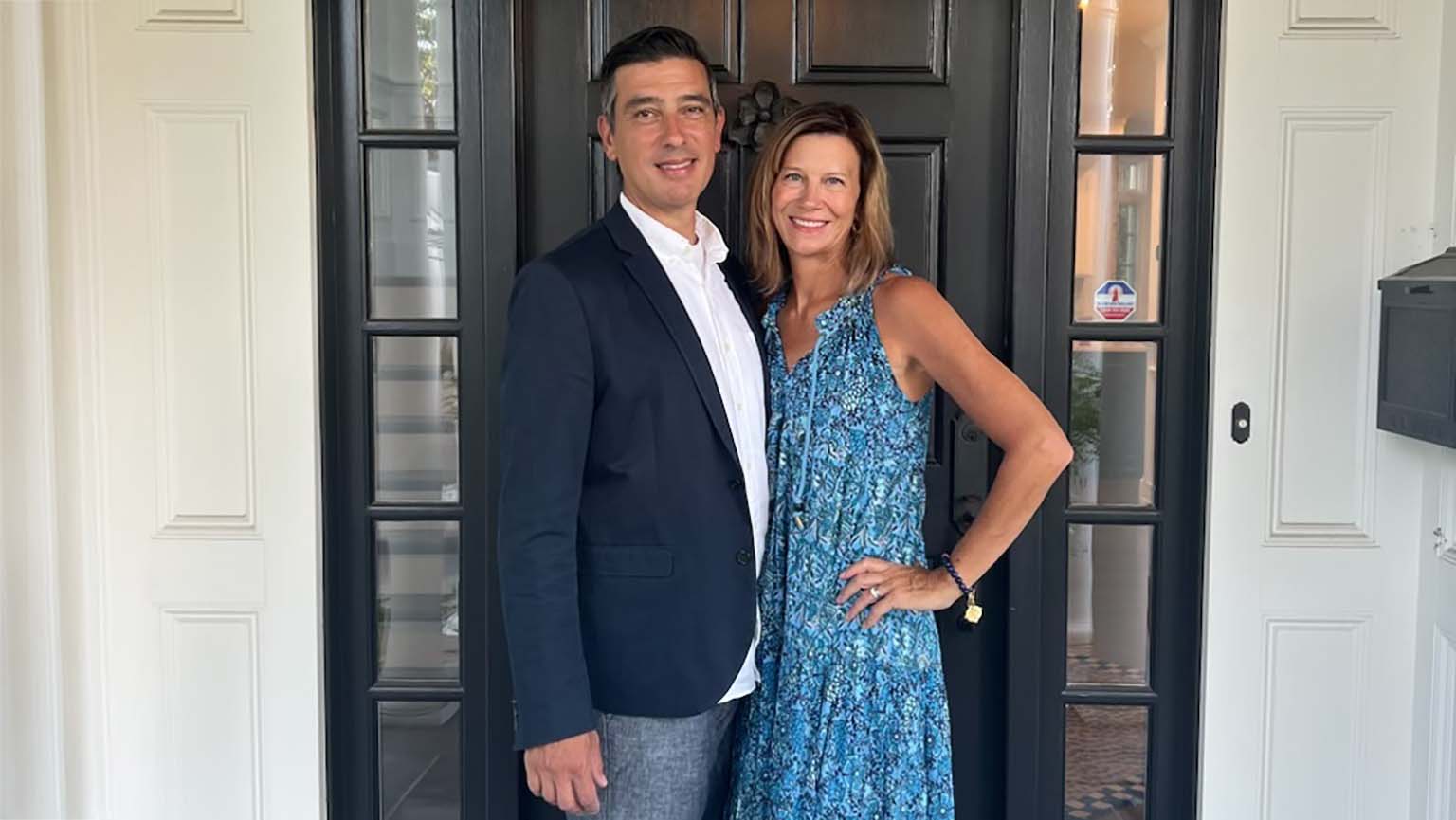 A man and woman stand together in front of a sleek black door, smiling at the camera.