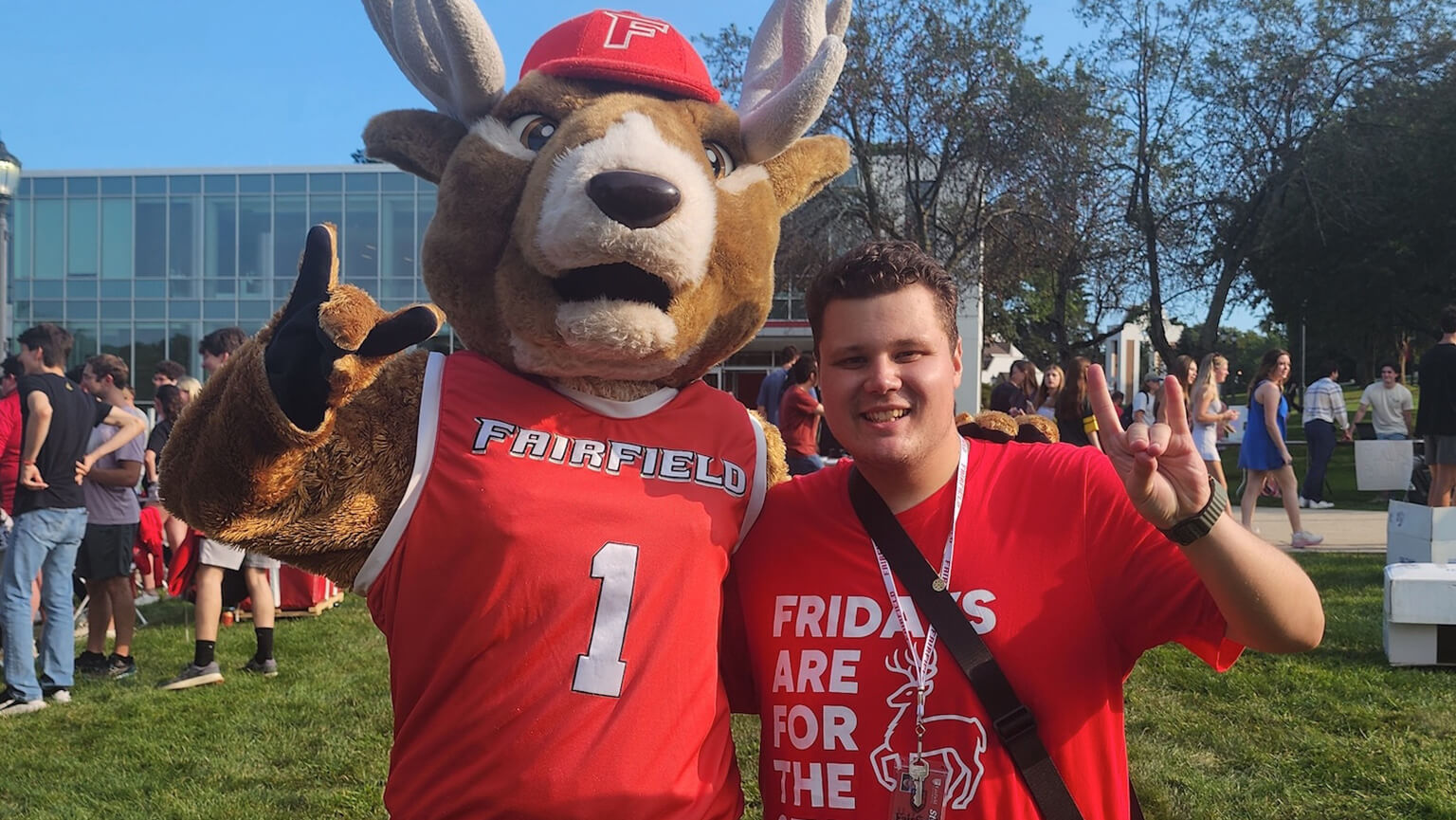 A student poses with Lucas the Stag outside on a bright sunny day on campus.